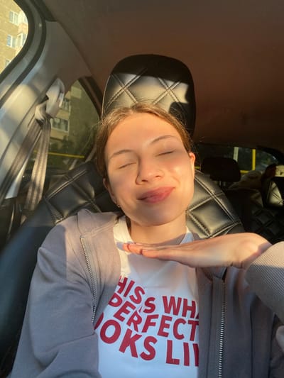 A young woman with her eyes closed is seated in the driver's seat of a car, bathed in warm sunlight. She is wearing a white t-shirt with red text that reads "THIS WHATEVER PERFECT LOOKS LIKE" and a light grey zip-up hoodie. Her right hand is gently resting on her chin. The car's interior features black leather seats with a diamond-stitched pattern. Through the car window, a building with multiple floors and windows is visible, suggesting an urban environment. The lighting indicates it is either sunrise or sunset.