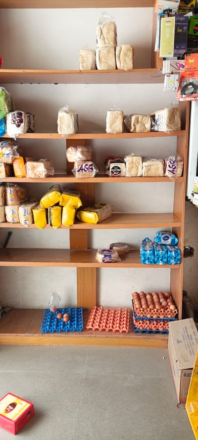 A wooden shelf displays assorted food items. The top shelf holds wrapped bread loaves and electronics. The next shelves down contain more wrapped bread, various brands visible. Underneath, there are City Fresh bread and wrapped yellow loaves. The subsequent shelf features City Fresh bread and packages of blue wrapped products. The bottom shelf showcases eggs in blue and orange containers, with some bare eggs and boxes on the floor.