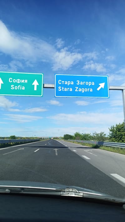 The image shows a view from a vehicle on a highway, likely in Bulgaria based on the road signs. Two prominent road signs hang overhead: one green sign points straight ahead towards "Sofia" (София), and a blue sign directs traffic to the right towards "Stara Zagora" (Стара Загора). The sky is blue with scattered clouds. The highway has multiple lanes with white directional arrows painted on the asphalt. The immediate foreground shows the vehicle's dashboard and windshield. The highway stretches into the distance, bordered by green vegetation on the sides.