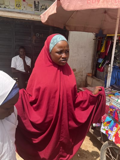 In Gura Top, Nigeria, a woman wearing a red hijab and a light blue headscarf is prominently featured in the foreground. She is looking to her right with a neutral expression. Behind her, a man in a white t-shirt and black pants is seated, with his hand on his chin. To the left, another person wearing a white hijab with a blue trim is partially visible, looking downwards. The scene appears to be an outdoor market, with stalls selling various items. In the background, there is a stall with clothing, including shirts with colorful skull prints and other patterns, as well as a blue jacket. An umbrella is positioned above the stalls, providing shade. The ground is dusty, and there is a visible portion of a wheel from a cart or bicycle on the right side of the image. A building with dark doors is visible behind the seated man, and a sign with text and phone numbers is mounted above it.