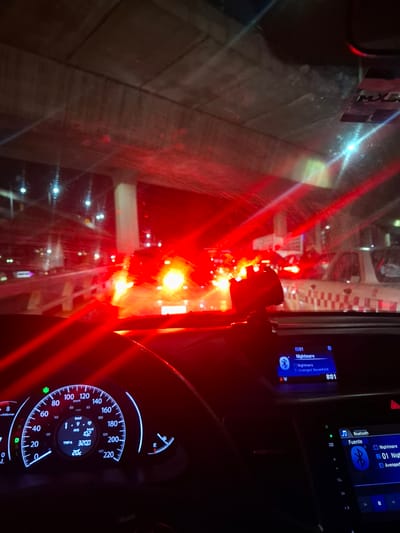 The image shows an interior view from the driver's seat of a car in Naucalpan de Juárez, Mexico. The dashboard is visible, displaying a speedometer with a reading around 60 km/h and a central console playing a song titled "Nightmare" by Avenged Sevenfold. Through the windshield, blurred red lights suggest a busy traffic scene. The reflection on the windshield obscures the precise details of the external environment. The car's windshield has a logo (MXG) on it.