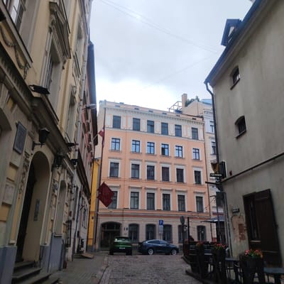 The image shows a narrow cobblestone street in Riga, Latvia, flanked by historic buildings. On the left, a tall, light-colored building with ornate architectural details and dark windows dominates the foreground. A gas lamp is visible on its facade. Further down the street, a pale peach-colored building with multiple stories and numerous windows is visible. Two maroon flags with a Latvian motif are hanging from the peach building. To the right, a lighter-colored building has a Guinness sign and a harp emblem. Outdoor seating with tables, chairs, and planters is set up on the sidewalk in front of this building. Two cars are parked on the cobblestone street; a green SUV on the left and a dark sedan on the right. The sky is overcast.