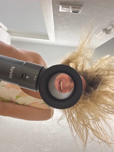 A low-angle, medium shot shows a woman holding a black hairdryer, with the nozzle pointed towards the camera. Her blonde hair is blown upward by the airflow, creating a wild, voluminous effect that fills the top of the frame. She is smiling broadly, with her mouth open, and appears to be enjoying the experience. The woman is wearing a light-colored sleeveless dress with a floral pattern. The background is a softly lit, pale textured wall, with some faint light fixtures visible on the left side of the image. The hairdryer has "tuvio" written on its body, along with a lock symbol and two triangle symbols. The overall impression is one of playful exuberance and carefree fun. The time of day and weather are not discernible from the image. The location is Votkinsk, Russia.