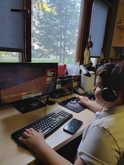 The image depicts a young boy wearing headphones, facing a computer screen, and playing a video game. He is seated at a desk with a keyboard, mousepad, and a smartphone. The computer screen displays a game with text and graphics, possibly a role-playing or simulation game. In the background, a window reveals trees and a building, suggesting an indoor setting possibly in the city of Марковский, Russia. The boy's posture and focus indicate engagement with the game. The overall scene portrays a typical gaming setup.