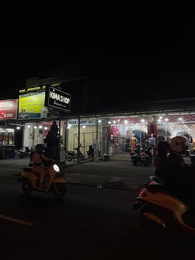 This is a night-time street-level photograph in Denpasar, Indonesia, featuring a commercial establishment with illuminated signs. The dominant business advertised is "KIMA SHOP," a clothing store, as indicated by the mannequins and clothing racks visible through its large glass storefront. Adjacent to KIMA SHOP is a laundry service, advertised with signs for "MESIN LAUNDRY" and "NON STOP LAUNDRY."

Several motorcycles are parked on the street or sidewalk, and one is in motion in the foreground, ridden by an individual whose face is obscured by a helmet. The street is paved and appears to be a busy area. The overall ambiance is that of a typical urban commercial strip at night.