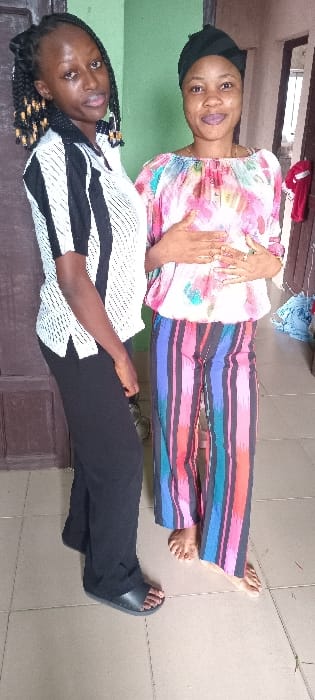 Two women are posing for a picture. The woman on top has on a pink and white top and colorful pants. She is wearing a black head covering. The woman on the bottom has on a white and black striped top and black pants. She is wearing black sandals. The women are inside a house with linoleum floors.