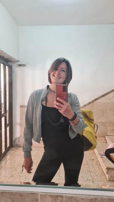 A woman takes a selfie in a mirror. She is wearing black pants, a black shirt, and a gray jacket. She is holding a red smartphone in her right hand. A yellow backpack rests on the floor behind her. The flooring is light brown with a speckled pattern. A person’s legs and feet are visible in the mirror’s reflection in the upper right corner. The wall on the left is white. The picture may have been taken in Nahariyya, Israel.
