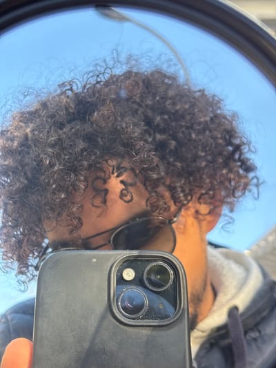 A male individual is depicted in a close-up selfie captured via a reflection in a circular or oval mirror, likely a vehicle's side-view mirror, in Tangier, Morocco. The subject has dark, densely curled hair and medium-brown skin, illuminated by bright daylight. He wears dark aviator-style sunglasses. A portion of a dark jacket with a lighter, possibly sherpa-lined, collar and a visible drawstring is present on his right side. He is holding a black smartphone with a triple-lens camera module and flash visible, possibly an iPhone, with his right hand, specifically his orange-tinted finger. The phone's camera lenses show minor dust particles. The reflection background includes a clear, bright blue sky and a slender, light-colored pole, possibly a street light, extending upwards against the sky.