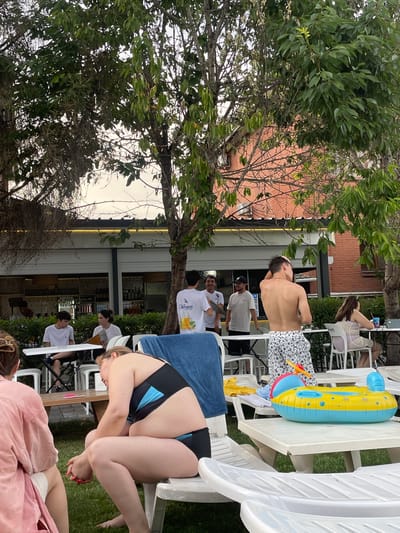 In this outdoor scene, possibly a poolside or garden setting in Almaty, Kazakhstan, a group of people are engaged in leisure activities. The time of day appears to be late afternoon or early evening, with soft, diffused lighting. Green trees and shrubs dominate the foreground and parts of the background, suggesting a warm, pleasant day. A brick building with windows is visible behind some of the foliage.

Several individuals are seated on white plastic chairs, some of which are arranged around tables. One person, a shirtless man with dark hair, is standing and touching his head. Another person, a woman wearing a black and blue bikini, is partially visible, leaning over. There are also other individuals in casual attire, including t-shirts and what appears to be a patterned dress. A yellow and blue inflatable ring toy rests on a white table. The presence of a blue towel also suggests a pool or water-related activity.

The atmosphere seems relaxed and casual, consistent with a day spent outdoors. The individuals appear to be socializing or simply enjoying their time. There are no overt signs of a specific event, but rather a general sense of people relaxing and enjoying the environment. The image is framed by the leaves and branches of trees, adding a natural element to the scene. The grass area visible suggests an outdoor space conducive to relaxation.