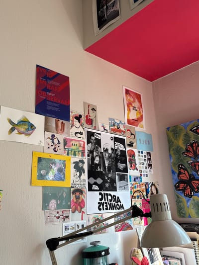 The image shows a section of a wall covered with various posters and artwork. The wall is painted white, except for a portion at the upper left which is a bright red. The posters include images of musical artists such as the Arctic Monkeys and Pulp Fiction, as well as various graphic designs and photographs. A desk lamp is positioned on the right side of the frame, its head pointing towards the wall. The scene appears to be in a room in Jesús del Monte, Mexico, based on the text "Jaffa" and "DITORA" and the local aesthetic. There are no people visible.