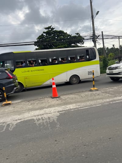 The image shows a busy street scene in Davao City, Philippines, with a yellow and white bus labeled "LOVE EXPRESS" prominently featured. The bus is traveling on a paved road, with several passengers visible through the windows. To the left of the bus, a dark-colored vehicle is partially visible, suggesting traffic. On the right side of the road, a white SUV with license plate "WAA 5998" is also in motion. The environment is characterized by overhead electrical wires, street lights, and lush green trees in the background, indicating an urban or suburban setting. A bright orange traffic cone is placed on the road, and a yellow and black striped traffic marker is visible near the left side of the frame, suggesting possible road work or a traffic diversion. The sky is overcast with gray clouds, typical of tropical weather.