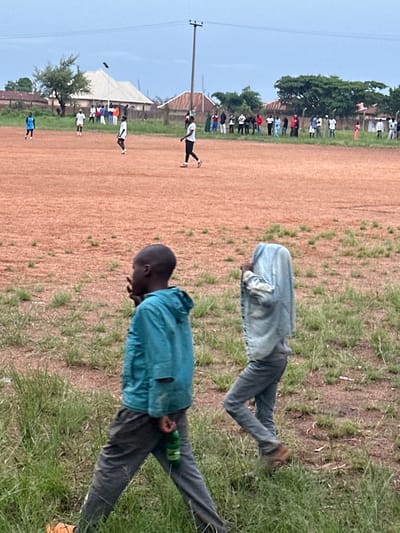The image depicts an outdoor scene, likely a sports field, on an overcast day. In the foreground, two children are walking on a grassy area with a dirt field behind them. The child on the left wears a teal hooded jacket and grey pants, with a green plastic bottle hanging from their pants. Their head is turned to the left, and their hands are raised in front of their face. The child on the right is largely obscured, with a light blue denim jacket draped over their head and upper body, their lower body clad in grey pants.

Further in the background, on the dirt field, several individuals are visible. Some appear to be playing or practicing a sport, while others are standing and observing. A group of people are gathered along the edge of the field. In the distance, buildings with pitched roofs and trees line the horizon. A prominent utility pole with wires stands to the right of the frame. The overall impression is of a community gathering for an outdoor activity in a semi-urban or rural setting. The presence of the utility pole suggests a connection to infrastructure. The reddish-brown color of the field indicates it is likely made of laterite soil, common in many parts of Nigeria. The cloudy sky suggests impending rain or a recent shower.