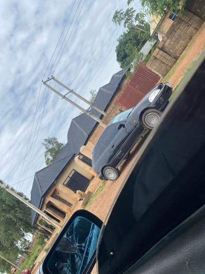 The image depicts a street scene in Kuru, Nigeria, as viewed from inside a car. The primary object is a black car parked on a dirt road. The car is a sedan with silver alloy wheels. The side mirror on the right side of the car is visible and appears to be cracked.
Behind the car, there is a house with a beige exterior and a dark grey corrugated roof. The house has several windows with dark frames. A power line pole with multiple wires stretches across the frame, indicating the presence of electricity infrastructure. Trees and vegetation are scattered around the scene, suggesting a semi-urban or rural environment. The ground is a mixture of dirt and grass.
The sky is cloudy, with a mix of white and grey clouds, suggesting an overcast day. The lighting appears to be diffused, consistent with cloudy weather. The orientation of the image is rotated, making it appear as if the scene is tilted. There is no visible text in the image. The overall impression is a mundane street view in a Nigerian locale, with the cracked side mirror being the most notable detail.
