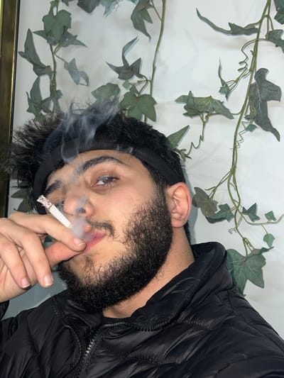 A bearded man wearing a black puffer jacket and head band is smoking a cigarette. He is looking into the camera and has light blue eyes. In the background are faux greenery that looks like ivy. The man is holding the cigarette in his fingers and there is smoke swirling around his face.