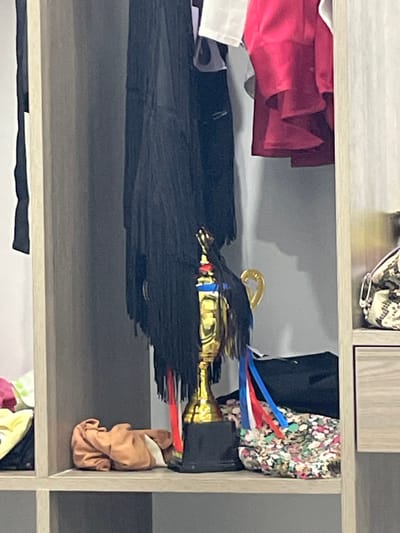 The image shows a closet with various items stored on shelves. In the center, a golden trophy with a black fringe and red, white, and blue ribbons is prominently displayed. To the left of the trophy, a black fringed garment hangs down. To the right of the trophy, a floral patterned fabric is visible. On the shelf below, a brown folded cloth is placed. In the background, red and white garments are hanging. The closet has wooden shelving and doors. The setting appears to be domestic. There is no discernible information about the city of Thuận An, Vietnam.