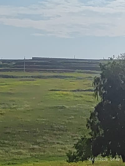 This is a landscape photograph taken on July 4, 2026, at 7:42 AM. The scene depicts a rural area with a large expanse of green fields under a bright, partly cloudy sky. In the background, there is a long, dark, terraced embankment or hill. A few tall, thin poles or markers are visible along the top of the embankment. On the right side of the image, a large, dark tree with dense foliage is prominent, partially obscuring the background. The image is taken from a distance, and the overall impression is one of an open, natural environment, likely in or near Konovalovo, Russia, given the timestamp and location context. There are no people or animals visible in the frame.