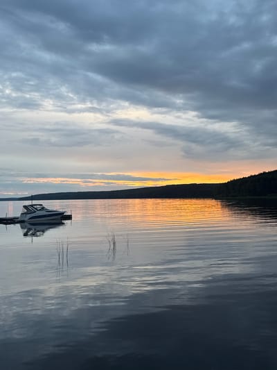 A low-angle, vertical photograph captures a serene lake scene at sunset in Chaykovsky, Russia. The sky is dramatically divided, with dark, brooding clouds on the left gradually giving way to a bright, warm band of orange and yellow light along the horizon on the right, indicating the setting sun. The water is calm and reflective, mirroring the sky and a dark treeline that runs along the far bank.
In the foreground, a small, white boat with blue stripes is docked. It is positioned on the right side of the frame and its reflection is clearly visible on the water's surface. The water itself is a deep blue-gray on the right side of the image, with lighter, rippled reflections of the sky and treeline on the left. The overall mood is peaceful and contemplative, enhanced by the soft light of the sunset. The image does not contain any visible text.