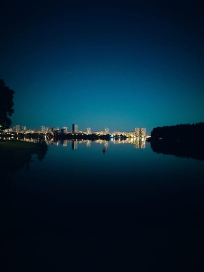 This is a nighttime photograph of the Svislach River with the skyline of Minsk, Belarus, in the background. The sky is a deep, clear blue, transitioning to a lighter blue near the horizon. A single, bright star is visible in the upper left quadrant of the sky.
The water of the Svislach River is dark and still, reflecting the lights of the city and the surrounding trees. On the left side of the riverbank, a silhouetted tree is partially visible. Across the river, a line of buildings, including high-rises and apartment complexes, are illuminated with numerous lights. The reflections of these lights shimmer on the water's surface, creating a distorted, elongated image.
On the right side of the frame, a dense line of trees forms a dark silhouette against the faintly lit sky. In the middle of the river, a single red buoy is visible, its reflection also appearing in the water. The overall impression is one of a peaceful, urban evening scene. There are no people or visible activity in the image.