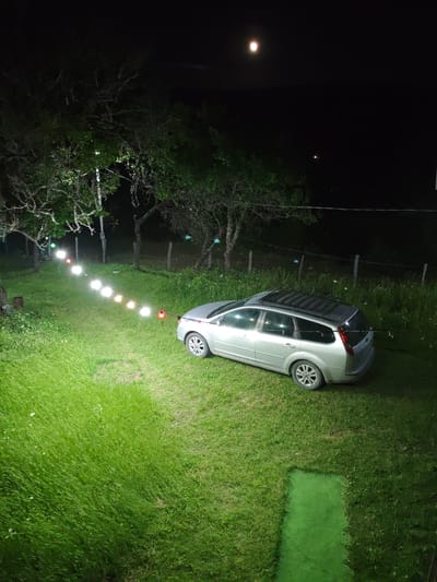 The image depicts a nighttime outdoor scene with a silver station wagon parked on a grassy area. String lights with multiple colored bulbs are strung across the yard, casting a warm glow. In the background, trees are visible against a dark sky where a bright moon is present. The setting appears to be rural or semi-rural, possibly a yard or a park. The location specified as Ukeshi, Georgia, is noted.
