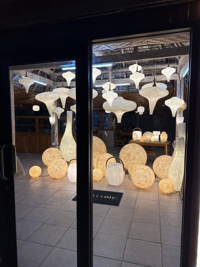 This media file is a photograph taken through a glass window, showing an interior scene. The image is oriented vertically, as if the viewer is looking down. The subjects are a collection of unique light fixtures, primarily lamps with organic, translucent shades. Some appear to be large, bell-shaped pendants hanging from the ceiling, while others are spherical and textured, resembling woven balls of light. These lamps are predominantly white and warm yellow, casting a soft glow on the surroundings.

The setting appears to be a retail space or a gallery. In the background, visible through the glass, are wooden architectural elements like beams and possibly shelving, suggesting a rustic or artisanal interior design. There are also reflections on the glass, including what looks like a doormat with the visible text "COME" on it. Further in the background, several Buddha head statues are visible on a shelf, adding to the unique decorative theme. The overall atmosphere is warm and inviting, likely taken at night or in a dimly lit environment, as the artificial lighting is the dominant source of illumination. The flooring is tiled. The location context provided is Banjar, Indonesia.