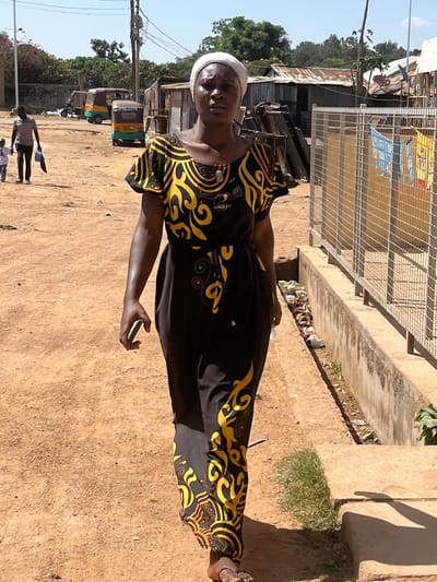 A woman wearing a black and yellow patterned dress and a white head covering stands on a dirt path in what appears to be Barakin Akawo, Nigeria. She holds a smartphone in her right hand. Behind her, there are a few parked auto-rickshaws, possibly a taxi stand. Some people are walking nearby. Above her is a pedestrian bridge over a drainage ditch filled with trash. The environment appears to be a busy commercial or residential area. Overhead powerlines and a satellite dish can be seen. The weather appears sunny. The picture is rotated 90 degrees counterclockwise.