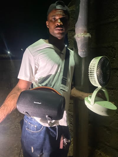 A man stands outdoors in Katsina Ala, Nigeria, lit by a hanging portable fan and light combination. He wears a white t-shirt, jeans, and a cap. A black speaker hangs across his body via a shoulder strap. An overhead pipe is visible in the background.