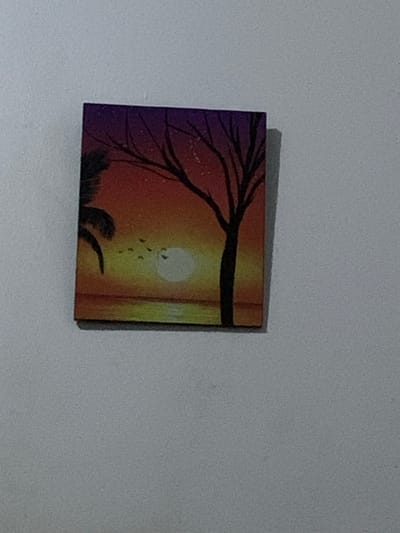A small, square painting of a sunset scene is affixed to a plain, light-colored wall. The painting depicts a dark silhouette of a tree with bare branches against a gradient sky of deep purple at the top, transitioning to orange and yellow towards the horizon. A bright, pale circle, representing the sun, is visible near the horizon. A few small, dark shapes in the sky suggest birds in flight. To the left of the tree, a dark silhouette of a palm frond is partially visible. The foreground of the painting shows a body of water with a reflective surface mirroring the colors of the sky. The wall is textured and appears to be a typical interior surface. The location in Barakin Lerawi, Nigeria, is not discernable from the image itself.