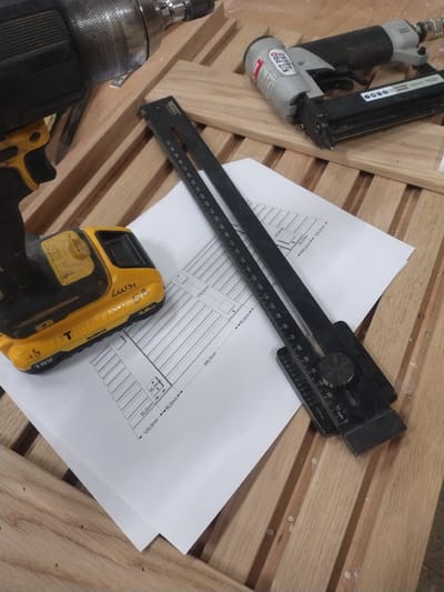 The image depicts a woodworking scene, likely in a workshop or garage. In the foreground, a yellow and black Dewalt power drill is positioned on a wooden surface composed of parallel planks. To the right of the drill, there is a white paper with technical drawings, displaying dimensions like "378.0mm," "345.0mm," "50.0mm," "20.0mm," "90.0mm," and "120.0mm." Overlapping the paper is a black marking gauge with a ruler and a circular adjustment knob. In the background, a gray and black pneumatic nail gun is placed on the wooden surface. The overall environment suggests a workspace where construction or furniture assembly is in progress. No specific landmarks of Leudelange, Luxembourg are visible, so the location cannot be determined from the image alone.
