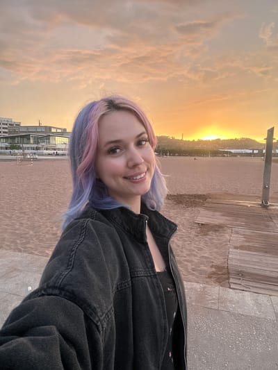A young woman with pink and blue dyed hair takes a selfie on a beach in Gijón, Spain. She is wearing a dark denim jacket over a black top. The background shows a sandy beach with a wooden boardwalk to the right. In the distance, there are buildings and hills, and the sun is setting, casting a warm orange glow across the sky. The overall scene is serene and depicts a casual moment at the beach.
