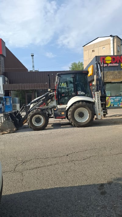 The image shows a white backhoe loader parked on an asphalt road in front of a building with a brown facade. The building has a prominent sign with red lettering that reads "RODN" and "Syn jurekten" in yellow. To the left of the building, there's a lighter-colored structure. The sky above is blue with scattered white clouds, suggesting it's daytime. The loader has "HIDROMEK" and "HHK 102b" written on its side. A website address "WWW.PCAGROUP.KZ" is also visible on the machine. The scene appears to be a commercial or industrial area. There are no people visible in the image. The overall impression is a bright, sunny day.