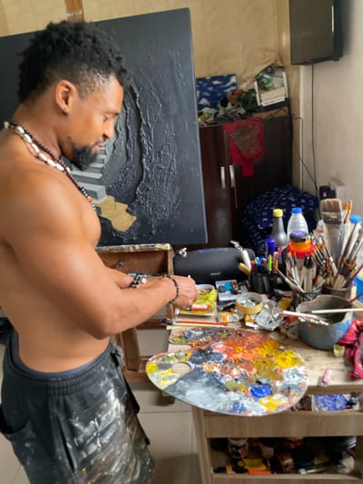 A bare-chested Black man with dark curly hair and a beard, wearing a necklace of beads and shells, and dark pants splattered with paint, stands in an art studio in Ado, Nigeria. He is looking down at a wooden palette filled with colorful paints, holding paintbrushes and a palette knife. To his left, a large black canvas with textured elements is propped up. The background shows a cluttered shelf with art supplies, including bottles of paint, jars of brushes, and other tools. The overall scene depicts an artist actively engaged in his creative process.