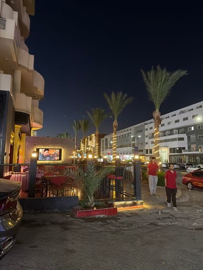 The image depicts an outdoor seating area of a restaurant or cafe at night, located in Hurghada, Egypt. The scene is illuminated by artificial lights, with decorative string lights wrapped around palm trees and lamps illuminating the walkway and seating areas. In the foreground, a dark-colored car is partially visible on the left. The seating area features pink sofas and red chairs arranged around tables. A large television screen mounted on a brick wall displays a man speaking. Two men wearing red shirts are walking away from the camera towards the right side of the image, along a paved walkway. Behind them, a modern, multi-story building with balconies and numerous windows serves as the backdrop, suggesting an urban or resort setting. The sky is dark, indicating it is nighttime.