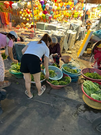 In Hội An Ward, Vietnam, a street vendor scene unfolds at dusk, illuminated by numerous colorful lanterns hanging overhead. A young woman in a pink shirt and patterned pants, wearing a white helmet, is seen from the side, bending down to examine produce. To her right, a woman in a white t-shirt and black shorts is also bent over, sorting through green vegetables in a basket. Behind them, an elderly woman in a plaid shirt and dark pants sits, seemingly engaged in packing or arranging items. The ground is paved with asphalt, showing signs of dampness and reflections from the lanterns. Various baskets filled with fresh green vegetables, including lettuce and leafy greens, are displayed on the ground. In the background, stacks of items covered in plastic sheeting and what appear to be suitcases are visible, suggesting a market or shop setting. A bright yellow and white sign stands vertically in the mid-ground, and a red fire hydrant with a yellow bucket nearby is present on the right side of the image. The overall atmosphere is that of a bustling evening market.