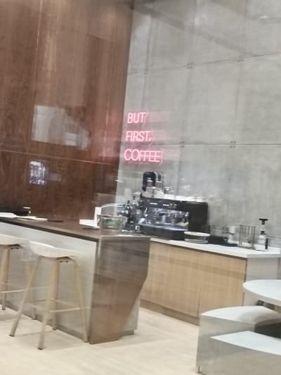 The image depicts the interior of a modern coffee shop. The setting is sleek and contemporary, with a concrete wall and wooden accents. A neon sign on the wall displays the text "BUT FIRST, COFFEE" in red, glowing letters, suggesting the primary purpose of the establishment.
Prominently featured is a professional espresso machine, indicating that this is a place where coffee is prepared with expertise. Various coffee-making accessories, such as pour-over devices, filters, and cups, are neatly arranged on the counter. The counter itself is a mix of stainless steel and wood, adding to the modern aesthetic.
In the foreground, there are white bar stools with thin, metallic legs, suggesting a seating area for patrons. The floor appears to be light-colored wood or laminate. To the right, a white table and seating area are partially visible, hinting at additional customer spaces.
There are no people visible in the image, giving it a quiet, perhaps early morning or late evening, atmosphere. The lighting appears to be interior, with the neon sign providing a distinct visual element. The overall impression is one of a well-equipped, stylish coffee shop in Dubai, United Arab Emirates, focused on providing a quality coffee experience.
