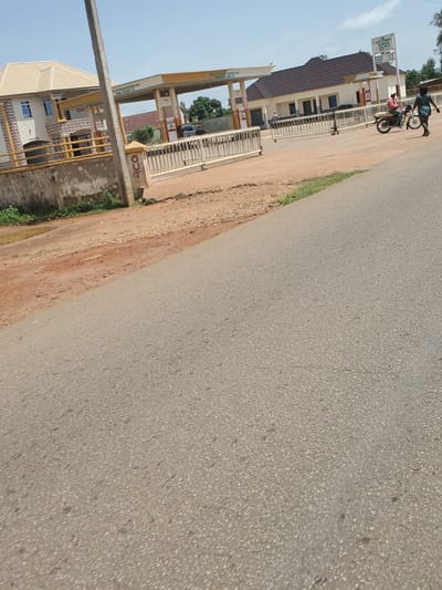 The image, taken in Vom, Nigeria, depicts a daytime roadside scene featuring a fuel station adjacent to a paved road under a clear, bright sky. The fuel station, prominently on the left, is identified by a yellow and brown canopy structure. A large vertical white sign displays the name "D. O. G. S. ENTERPRISES LIMITED" and advertises "Quality ASSURED QUANTITY," along with "OUR CORE VALUES" listed as Safety, Integrity, Customer Focus, Excellence, and Teamwork. Another partial sign on a canopy support structure reads "D. O. G. S. ENERGY RESOURCES AND INVESTMENT LIMITED."

Beneath the canopy, several fuel pumps are visible, indicating "PMS" (Premium Motor Spirit) and "DIESEL," with "N0.00" displayed for prices. A building with a brown roof and light-colored walls stands behind the pumps, separated by a white metal fence or railing. Two individuals are near the fuel station's entrance area. One person, wearing a dark shirt and dark trousers, is standing next to a motorcycle or scooter. The second individual, dressed in a red top and dark trousers, is seated on the same motorcycle. Their activity suggests a stop at the fuel station, possibly for refueling or a break.

The road surface in the foreground and stretching into the distance is grey asphalt. Alongside the road is a wide, unpaved shoulder of reddish-brown dirt and gravel, which transitions into patches of green grass and vegetation. A grey utility pole stands upright in the lower-left foreground. A concrete pillar or block with the letters "JCG" is also visible near the roadside. The bright lighting indicates the time of day is likely late morning or afternoon.
