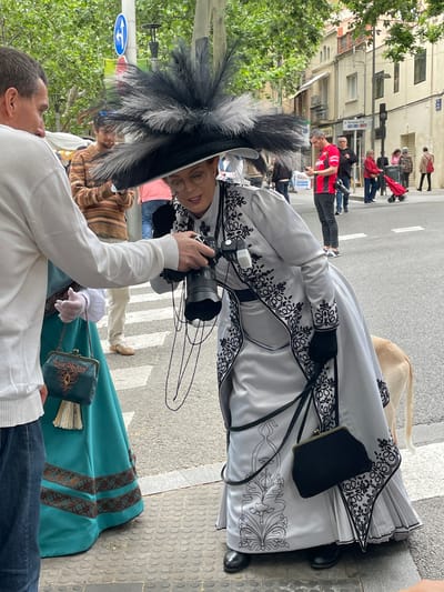 The image depicts a street scene in Terrassa, Spain, with pedestrians walking in the background. In the foreground, two individuals are interacting. One person, likely a photographer, is holding a Sony camera and is poised to take a picture. The other person is wearing a striking white and black period costume with elaborate embroidery and a large feathered hat, a small dark purse, and a long pleated skirt. A fan is visible near them. A blue and green dress of a similar period is slightly visible. The interaction takes place on a pedestrian crossing, and the surroundings include buildings and greenery, typical of an urban setting in Terrassa.