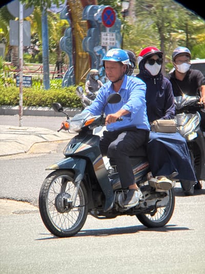 In Nha Trang, Vietnam, three individuals are traveling on two motorcycles. The primary motorcycle in the foreground is ridden by a male driver wearing a blue helmet and a light blue collared shirt. He is followed by a female passenger wearing a red helmet, dark sunglasses, and a patterned face mask. The second motorcycle behind them has a male driver wearing a gray helmet and a dark shirt, and another passenger is partially visible. The people are on a street, with a sidewalk and greenery in the background. The lighting suggests it is daytime.