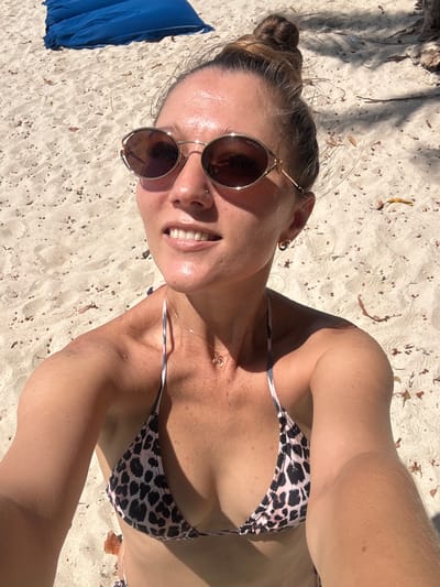A woman is taking a selfie on a sandy beach in Baan Tai, Thailand. She is wearing a leopard print bikini and sunglasses. The sand is light-colored with scattered leaves and debris. In the background, a large blue object, possibly a cushion or mattress, is visible on the sand. The lighting suggests it is a sunny day, with shadows cast across the sand.