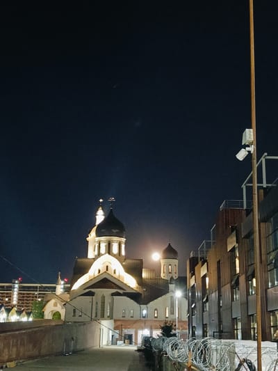 The image shows a nighttime view of a church with a prominent dome, illuminated by lights. The church appears to be in the city of Krasnodar, Russia, possibly at the Pokrovsky Cathedral. A large moon is visible in the dark sky, near the church dome. To the right of the church, there is a modern building with many windows. A tall pole with security cameras is also visible. In the foreground, there is a brick wall and barbed wire fencing. The overall scene suggests an urban environment at night, with a blend of historical and modern architecture.