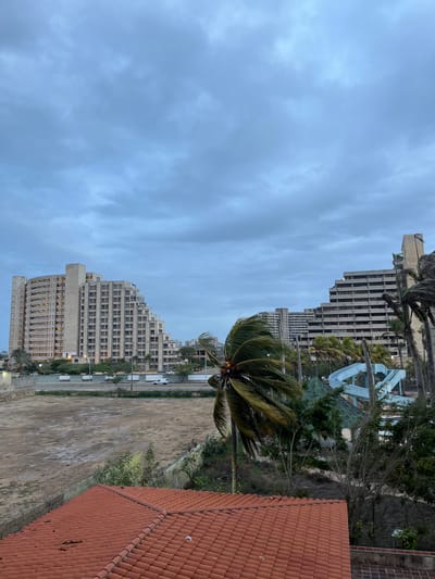 The image captures an outdoor scene in Porlamar, Venezuela, on a cloudy day. In the foreground, a reddish-tiled roof is visible. Beyond the roof, a large, empty, dirt lot takes up a significant portion of the mid-ground. To the left of the lot, a modern, multi-story building with a unique stepped architectural design and numerous windows stands out. To the right of the lot, another similar building is visible, partially obscured by palm trees. In the background, a light blue water slide can be seen amidst dense greenery. Several palm trees are scattered throughout the scene, with one in the mid-ground showing fronds being blown by the wind. A road with a white vehicle driving on it runs along the edge of the dirt lot, separating it from the buildings. The sky is overcast with gray clouds.