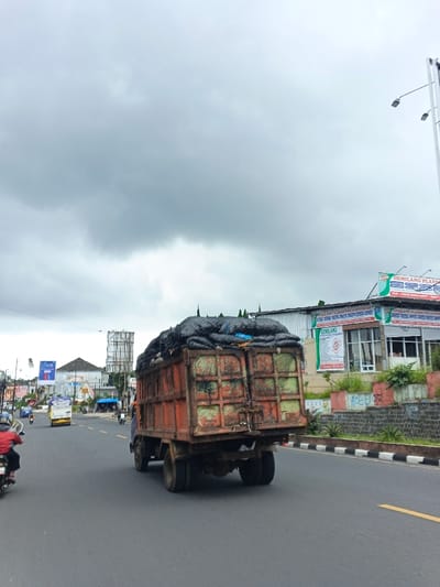 The scene depicts a daytime urban street in the city of Palayan, Philippines, under a heavily overcast sky. An old, rust-colored cargo truck with dual rear wheels, heavily loaded with dark material covered by a black net/tarp, is the prominent subject, traveling away from the viewer on an asphalt road. The truck exhibits significant wear and rust on its body.
To the immediate left, a red motorcycle with a rider wearing a red jacket is partially visible, also moving away. Further down the road on the left, other vehicles, including another motorcycle and a white utility van, are present. The road features a solid yellow centerline, white dashed lane markings, and a black and white striped concrete curb/median on the right.
On the right side of the road, a two-story commercial building with a light-colored facade and large windows is visible. A prominent green and white sign on its upper level reads "GEMILANG PLATON PVC INTERIOR" along with contact information. A low retaining wall with some vegetation and faded graffiti sits in front of the building. Tall streetlights with modern fixtures line this side of the road. In the distance on the left, various commercial structures, including a building with a "Candy" sign and a large billboard structure, are discernible. Power lines traverse the upper portion of the frame.