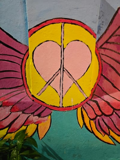 This image displays a vibrant mural painted on a wall in Kochi, India. The central artwork consists of a yellow circular emblem outlined in red. Within this yellow circle, a pink heart shape is prominently featured, bisected by the black lines forming a peace symbol. Stylized wings, rendered in shades of pink, purple, and orange, extend outwards from both sides of the central circular emblem. The upper section of the wall is painted light blue, while the lower section is teal. In the bottom left corner, green foliage from a plant is visible, partially obscuring the base of the left wing and the teal wall.
