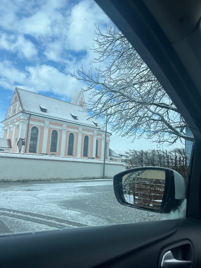 A view from inside a vehicle looking through the driver's side window towards a church building in Benningen, Germany. The church has a white and pink facade with a snow-covered roof. Bare tree branches are visible in the sky above the church. The side mirror of the car reflects a building and a snow-covered street. The road appears to be icy or covered in snow.