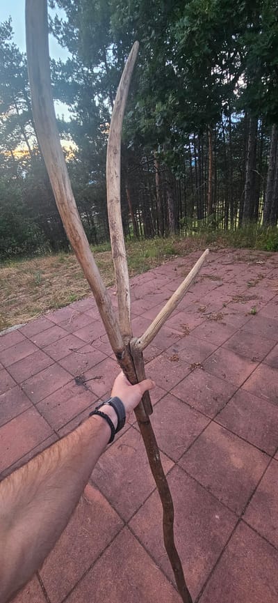 In Momin Prohod, Bulgaria, an individual's forearm, adorned with a dark grey wristband and a black braided bracelet, is visible holding a roughly assembled wooden tool. The tool is constructed from three weathered branches, joined together at a central point with what appears to be wire or twine. Two branches angle upwards and outwards, while a third, shorter branch extends to the right. The tool's main handle is a longer, curving branch that extends downwards. The ground is paved with red, diamond-shaped tiles. Behind the paved area, dry grass and sparse vegetation lead to a dense forest of pine trees. A green hammock is visible suspended between trees in the background. The sky appears to be a pale blue with hints of sunset colors.