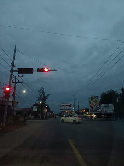 This image depicts a street scene at dusk in Manyang Cut, Indonesia. The sky is overcast with dark gray clouds. A traffic light on the left side of the road is illuminated red. Another traffic light, mounted on a horizontal pole extending over the road, is also showing a red light. Power lines crisscross the sky, supported by utility poles. A white sedan is visible driving away from the camera on the right side of the road. In the background, there are buildings and several large billboards, one of which displays the number "76" and the word "DARUM" and "SEGER BIKHE". The road itself is asphalt, with a yellow dividing line visible in the foreground. The overall impression is one of twilight in an urban or semi-urban environment.