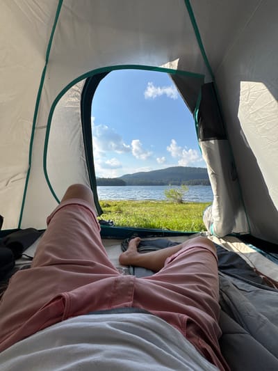 An outdoor, eye-level, medium shot captures the interior of a tent with a person's legs in the foreground and a scenic view of a lake and forest through the tent opening. The person is wearing pink shorts and a white shirt. Their legs are bent, with one foot visible, wearing a sandal. Inside the tent, a sleeping bag is laid out. Outside the tent, a grassy area leads to a calm lake, with a densely forested hill in the background under a bright blue sky with scattered white clouds. The sunlight casts shadows on the tent's interior. The scene is likely located near Sarnitsa, Bulgaria, given the natural landscape.