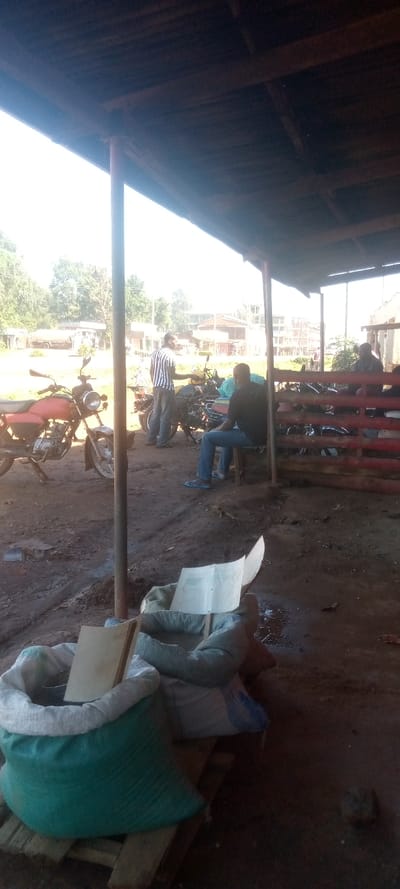 The scene is taken under a corrugated iron roof in a village in Kivaywa, Kenya. The ground is dusty. There is a man in a black and white shirt, he appears to be interacting with a man sitting on a chair. The man sitting on the chair is wearing a blue shirt, jeans and sandals. There are numerous motorbikes in the scene. There is a large green bag with what looks like a white book on top of it, and a white bag next to it.  There is a small piece of paper on the ground. The image was likely taken from a slightly elevated angle. The location is close to the coordinates 0.56, 34.806.  The scene shows a typical gathering place for men in a rural Kenyan community.