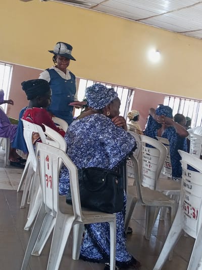 The image appears to be taken indoors, possibly a meeting hall or church, in Zango-ndaji, Nigeria. Several women are seated in white plastic chairs. Most are wearing patterned blue and white clothing and head coverings. A woman in a blue dress and a hat is standing and handing out small items, likely pamphlets or cards, to the seated women. The hall has light-colored walls, windows with bars, and a tiled floor. Some of the chairs have numbers or letters on them, and the fluorescent lighting is visible on the ceiling.