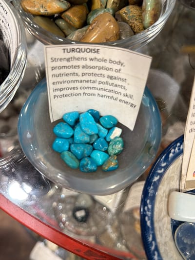 The image displays a close-up, slightly overhead view of a retail display featuring various polished stones, likely within a glass display case or on a reflective counter. At the center is a small, transparent bowl with a light-blue hue, filled with numerous tumbled turquoise stones. These stones exhibit a range of blue and greenish-blue tones, with some showing natural brown or white matrix patterns. Behind this bowl, a white paper label identifies the stones as "TURQUOISE" and lists their purported benefits: "Strengthens whole body, promotes absorption of nutrients, protects against environmanetal pollutants, improves communication skill. Protection from harmful energy."

To the upper left, a portion of another clear bowl is visible, containing larger, irregularly shaped, tumbled stones that are predominantly tan and brown with some greenish areas. A partial label above this bowl shows "ONYX" and mentions "stancy", "Self-Con", and "Vigor". To the right of the turquoise bowl, a blue and white patterned ceramic plate or dish is partially seen, holding a smooth, white, rectangular object and a translucent, blue, heart-shaped object.

The reflective surface beneath the items shows blurred reflections of the surrounding environment, including light sources and indistinct forms. A red frame, consistent with the edge of a display case, is visible in the bottom right corner. No individuals are present in the image. The scene is consistent with a metaphysical shop, crystal store, or gift shop. The specific location within New York City, United States, is not identifiable from the visual information provided.