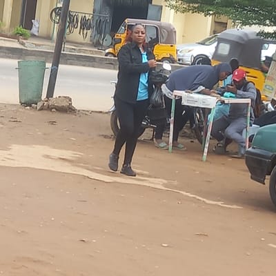 The image captures a street scene in Dadi Riba, Nigeria. The foreground shows a young woman wearing a black jacket over a blue top and black leggings standing on the sidewalk. She is looking directly at the camera. Behind her are several motorized tricycle taxis (Keke Napeps) parked along the side of the road. A group of people, including a woman in a hijab and others possibly studying or working at a makeshift table, are gathered near the tricycles. The background includes buildings and street infrastructure like poles and bicycle racks. The road and sidewalk are primarily dirt and show signs of wear.